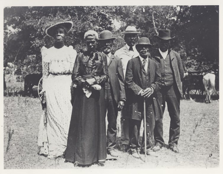 A group of people in fancy dress from the turn of the 19th century, standing together outside.
